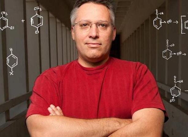 headshot of delmar larsen in a red shirt in front of a chalkboard with chemistry structures 