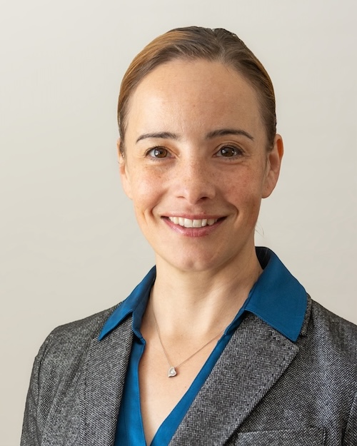 Professional headshot of a woman with blonde hair pulled back in a ponytail wearing a blue collared shirt and gray suit jacket