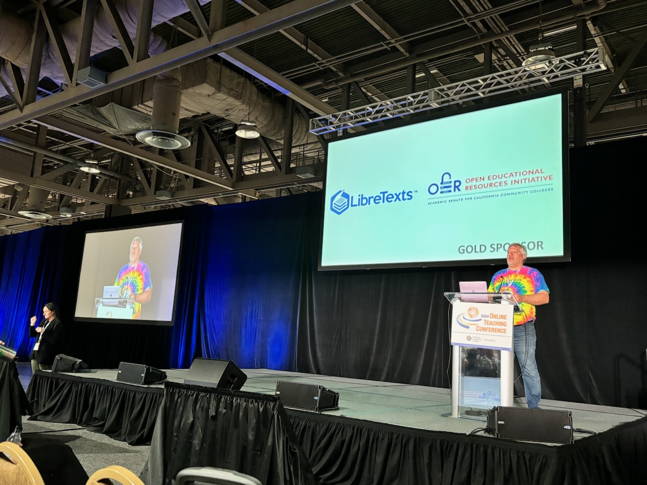man behind podium on stage in front of screen with logos for LibreTexts and OERI
