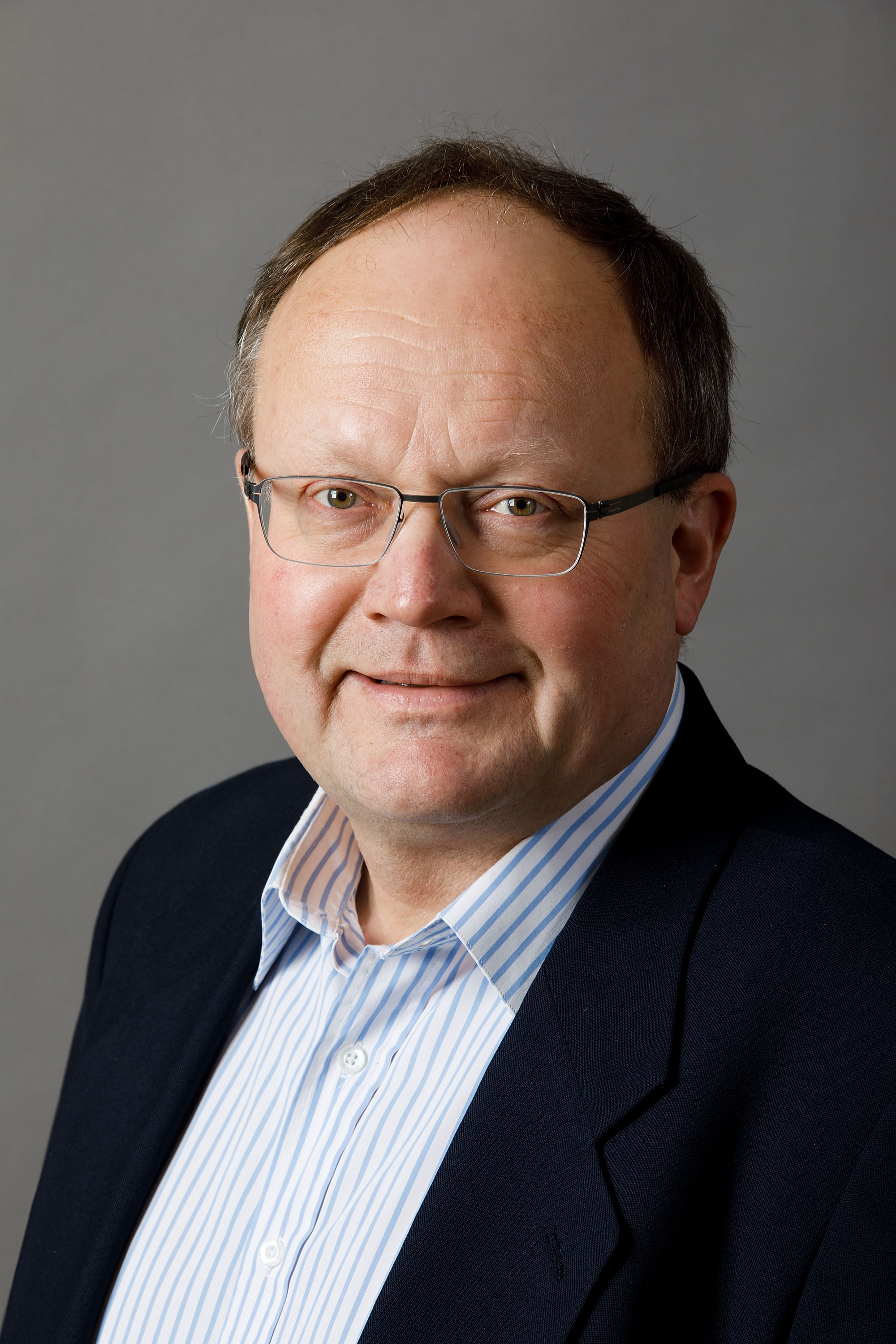 professional photo of a man in a black suit jacket, light blue shirt, and glasses smiling for the camera in front of a gray backdrop