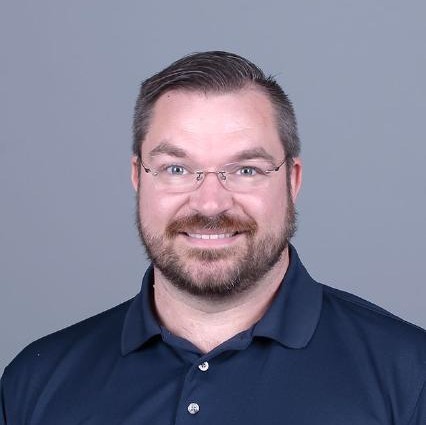 professional photo of man with glasses and beard standing in front of gray background smiling for the camera