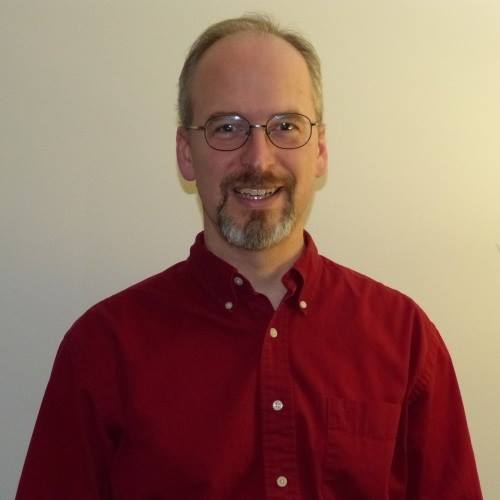 man in a red button down shirt and glasses smiling for the camera in front of an off white background