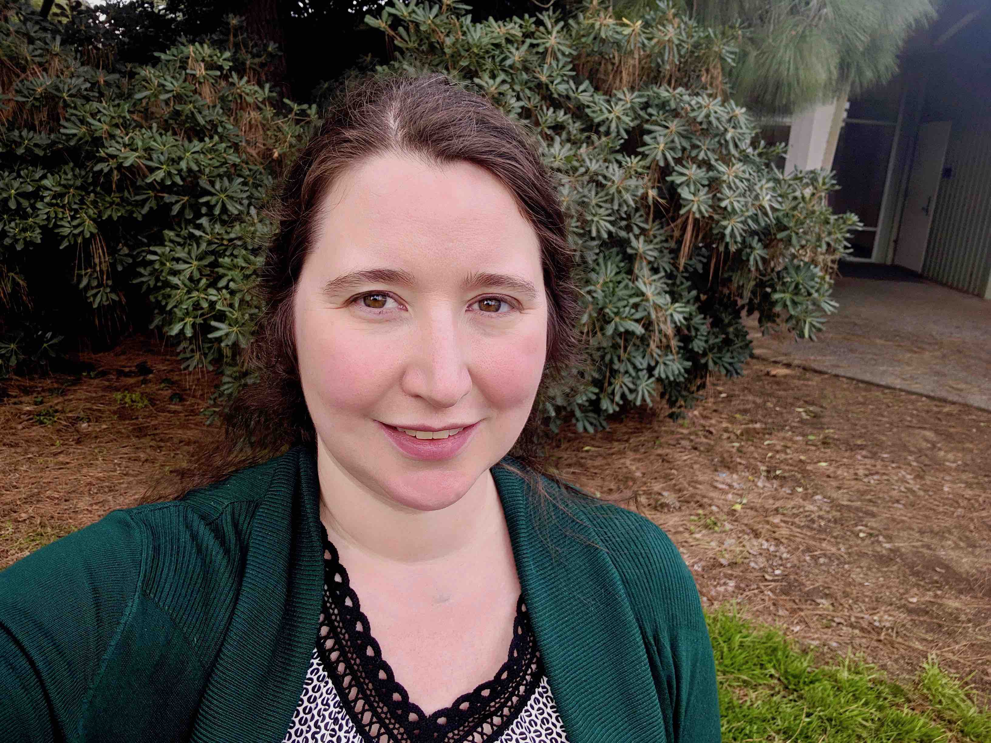 woman in a green sweater smiling for the camera while standing in front of some bushes