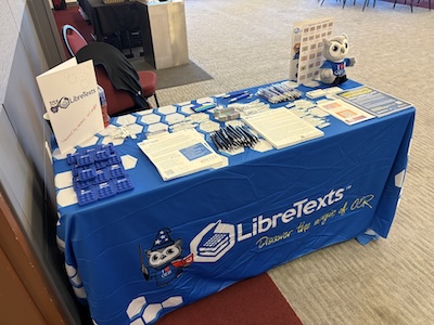 picture of a conference booth with various swag items on top and a blue tablecloth with LibreTexts wordmark in white.