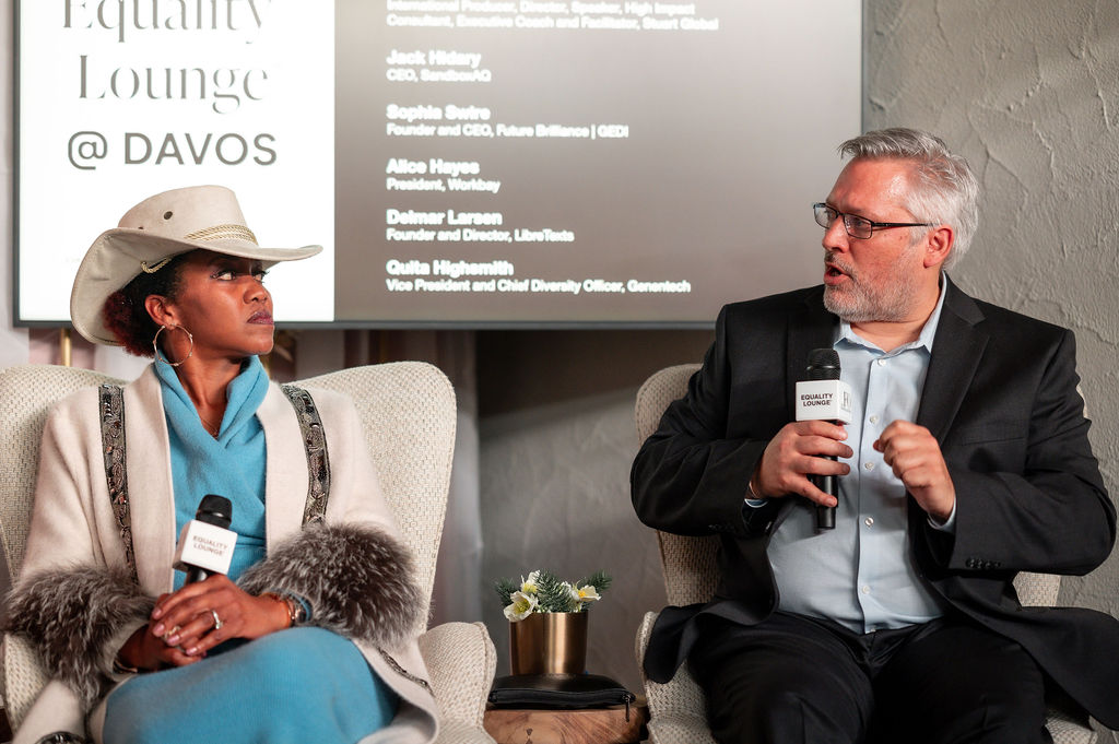 White man in a suit and glasses (Founder, Delmar Larsen) sitting on a stage identified as @Davos conference chatting with a black woman in teal and white outfit with white cowboy hat. 